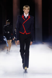 Runway model wearing a navy blazer with bold red piping, white shirt, black trousers, and yellow socks walking through fog on the catwalk.