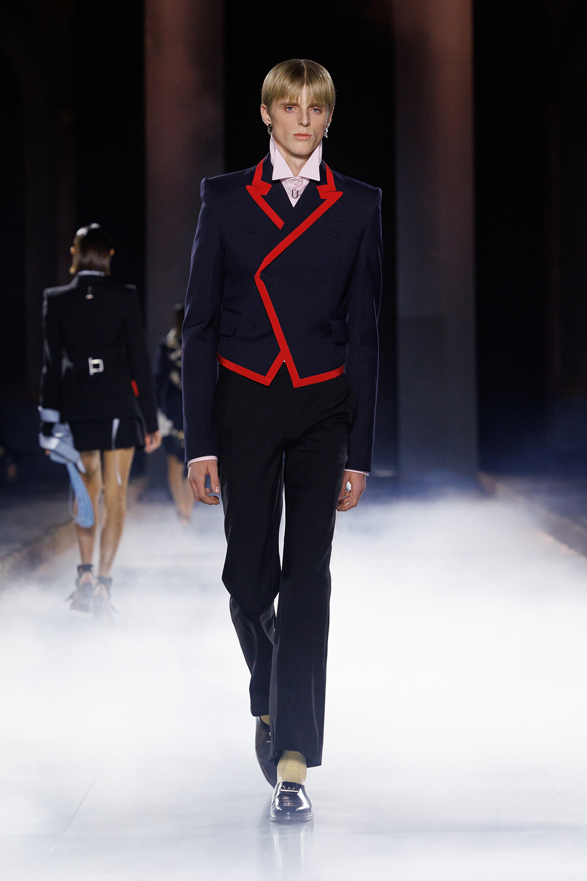 Runway model wearing a navy blazer with bold red piping, white shirt, black trousers, and yellow socks walking through fog on the catwalk.