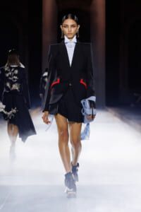 Model walking the runway in a black structured blazer with red trim pockets, a white shirt, and black shorts, holding a light blue handbag with fringe details amid a foggy fashion-show backdrop.