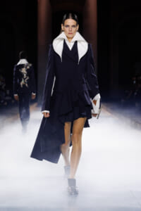 Model on a fashion runway wearing a navy double-breasted coat with an oversized white collar, a layered skirt, and a white handbag amid foggy lighting.