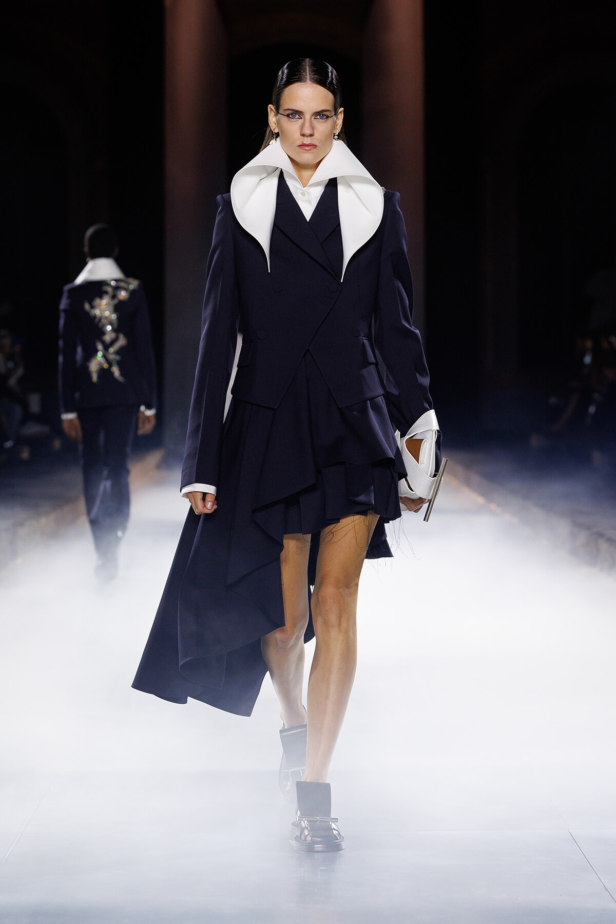 Model on a fashion runway wearing a navy double-breasted coat with an oversized white collar, a layered skirt, and a white handbag amid foggy lighting.