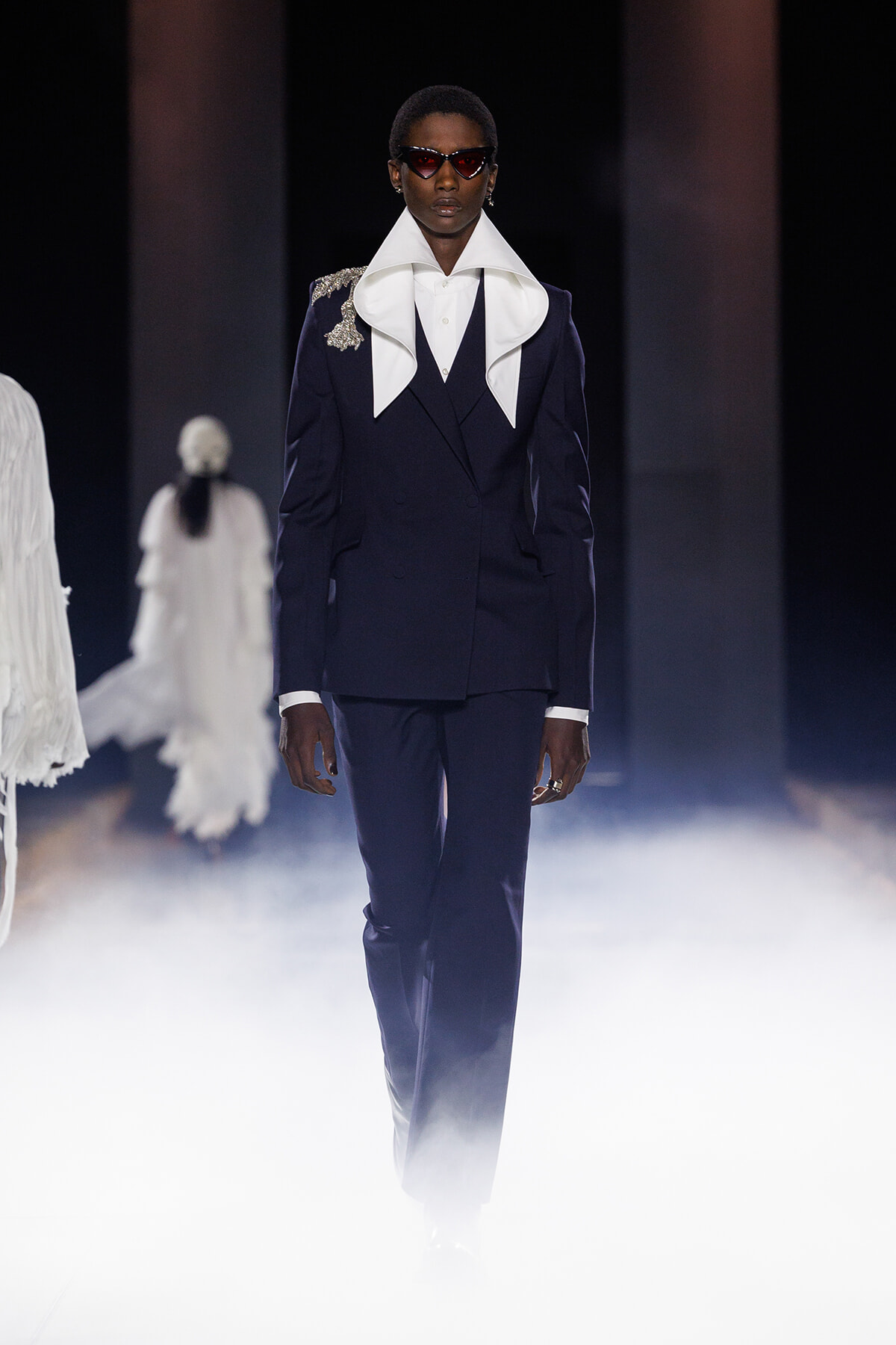 Model in a navy double-breasted suit with oversized white lapels and a decorative shoulder detail, wearing red sunglasses, walking through fog on the runway.