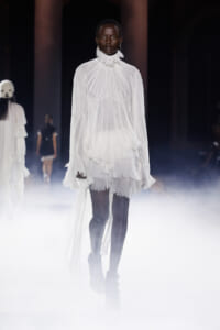 Model walking the runway in a dramatic white pleated high-neck dress with a long trailing cape, foggy stage lighting in the background