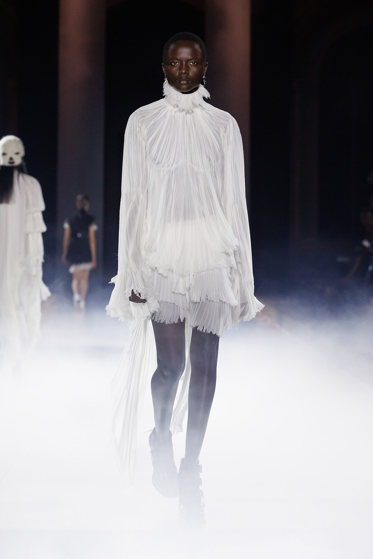 Model walking the runway in a dramatic white pleated high-neck dress with a long trailing cape, foggy stage lighting in the background