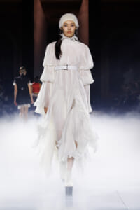 Model walking a runway in a voluminous white pleated dress with layered sleeves and a white belt, surrounded by fog.