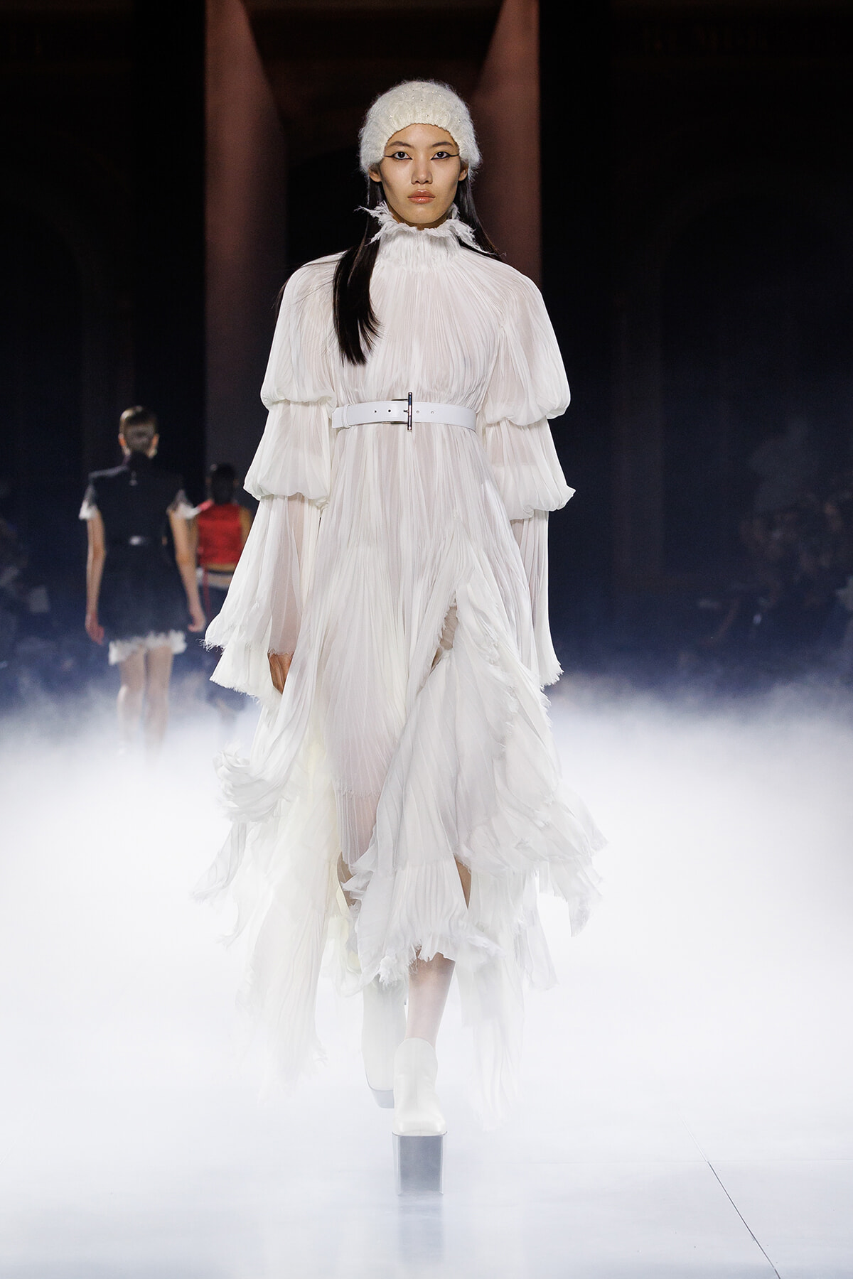 Model walking a runway in a voluminous white pleated dress with layered sleeves and a white belt, surrounded by fog.