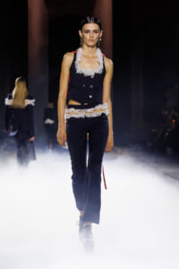 Model on a runway wearing a navy vest with white frilled lace accents and matching navy pants, surrounded by fog.