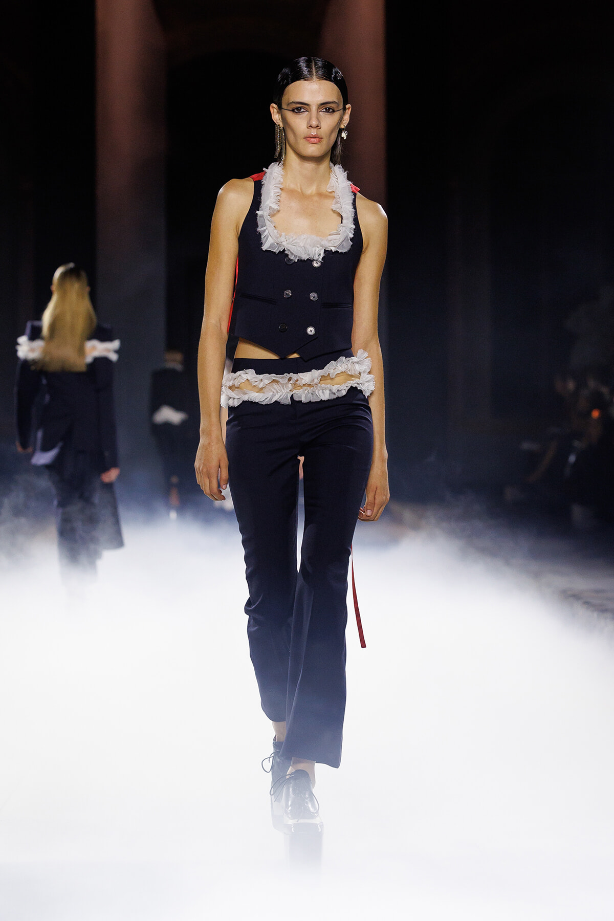 Model on a runway wearing a navy vest with white frilled lace accents and matching navy pants, surrounded by fog.