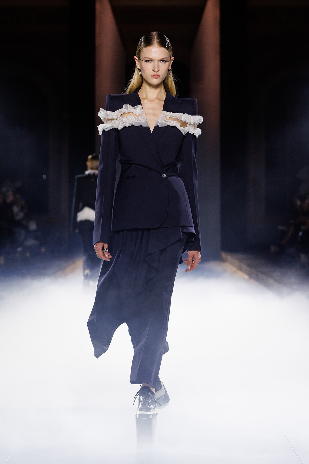 Model walking a runway in a navy tailored blazer with a wrapped, layered skirt and white fringe along the shoulders; smoky, spotlighted catwalk.