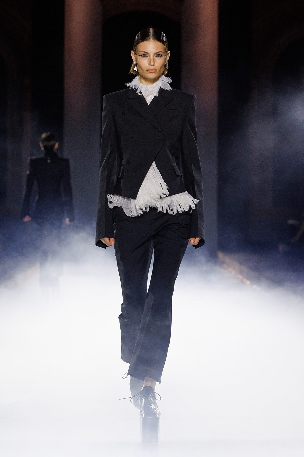 Model in a black double-breasted suit with white fringe at the collar and hem, walking through fog on a runway.