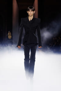 Model walking a dark runway in a structured black suit with dramatic shoulders, surrounded by fog and low lighting.
