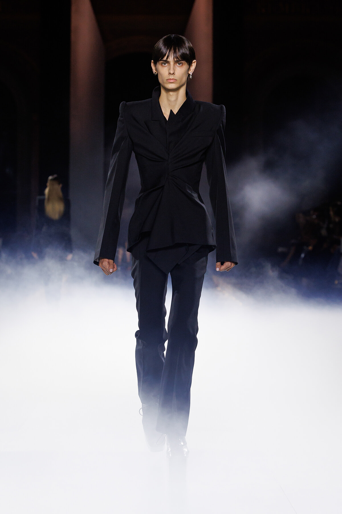 Model walking a dark runway in a structured black suit with dramatic shoulders, surrounded by fog and low lighting.