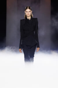 Female fashion model in a structured black blazer and matching pants walking down a smoky runway.