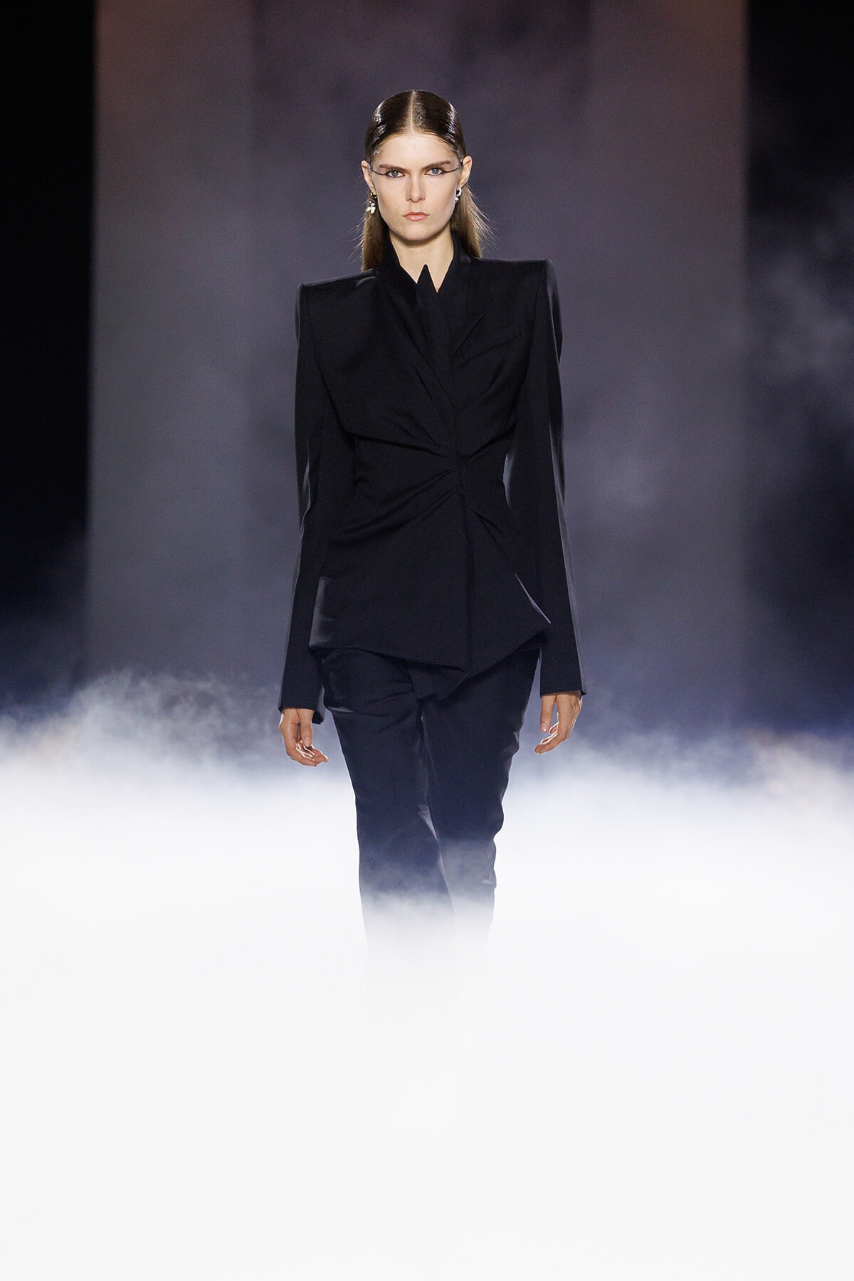 Female fashion model in a structured black blazer and matching pants walking down a smoky runway.