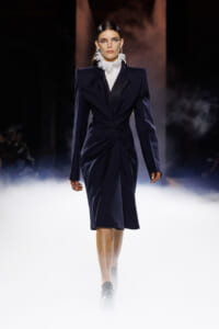 Model walking a dark navy, structured knee-length dress with a cinched waist and bold lapels, white ruffled high collar, on a foggy runway.