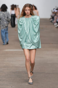 Model walking a runway in a metallic mint-green draped mini dress with exaggerated shoulders and gathered neckline, wearing gray strappy heels.