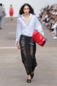 Model walks the runway in an oversized light blue button-down shirt and a black shredded knit skirt, carrying a bold red bag.