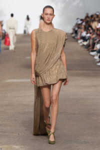 Model walking runway in a gold sequined draped mini dress with a long train on one side and olive green sandals.
