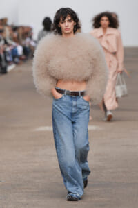 Model on a runway wearing a voluminous beige fluffy cropped sweater paired with high-waisted loose blue jeans and a black belt, audience blurred in the background.