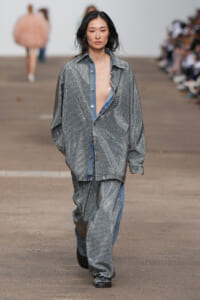 Model walking runway in oversized silver sequin suit with blue denim accents, neutral expression, hands in pockets.