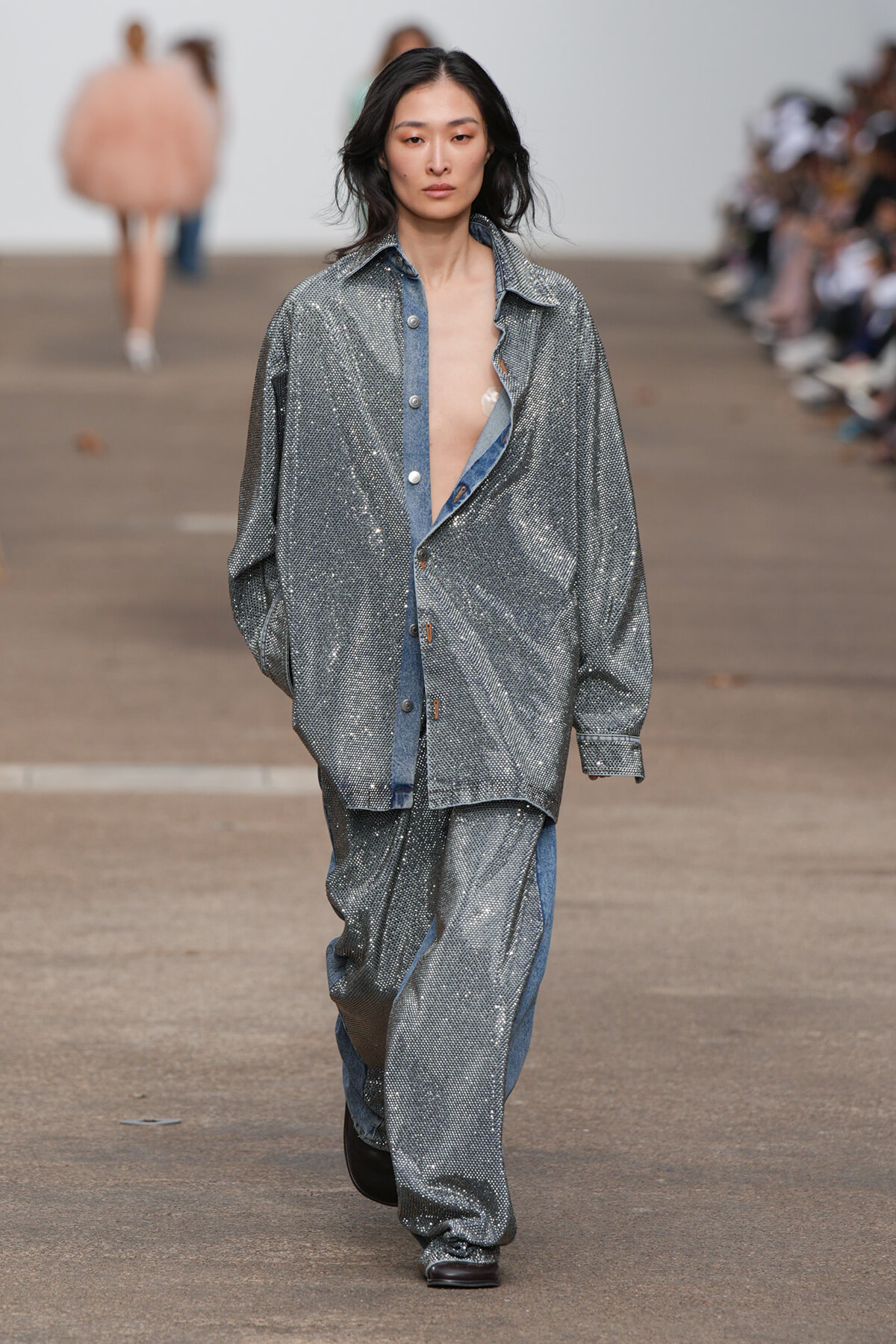 Model walking runway in oversized silver sequin suit with blue denim accents, neutral expression, hands in pockets.