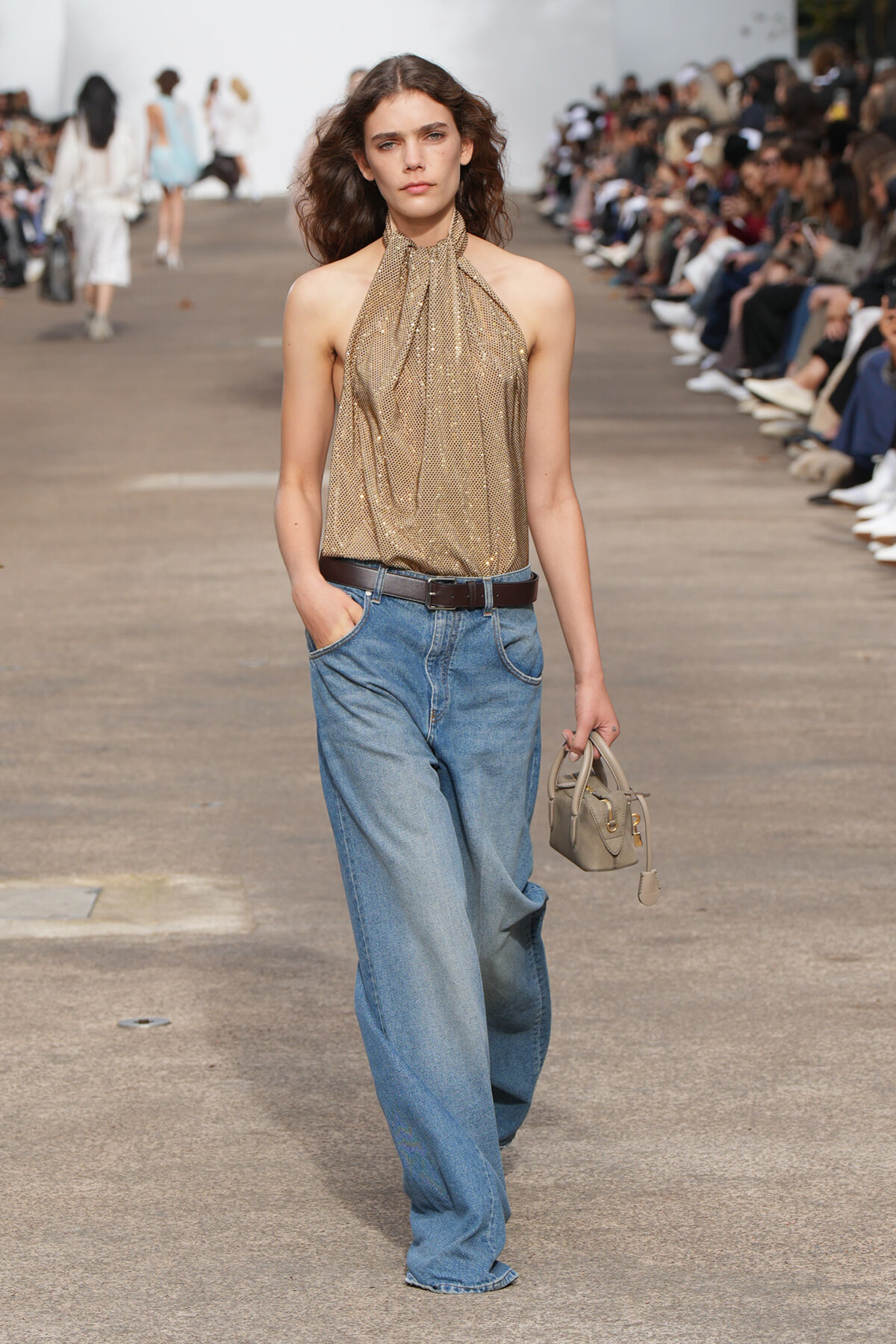 Model walks runway in a gold halter top and wide-leg jeans, holding a small beige handbag; audience lines the catwalk.