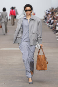 Model walking a runway in a light gray jumpsuit with a gray jacket, black sunglasses, and a tan brown tote bag.