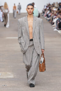 Model walking the runway in an oversized gray beaded pinstripe suit, blazer open and shirtless, carrying a brown tote with newspapers inside at the side.