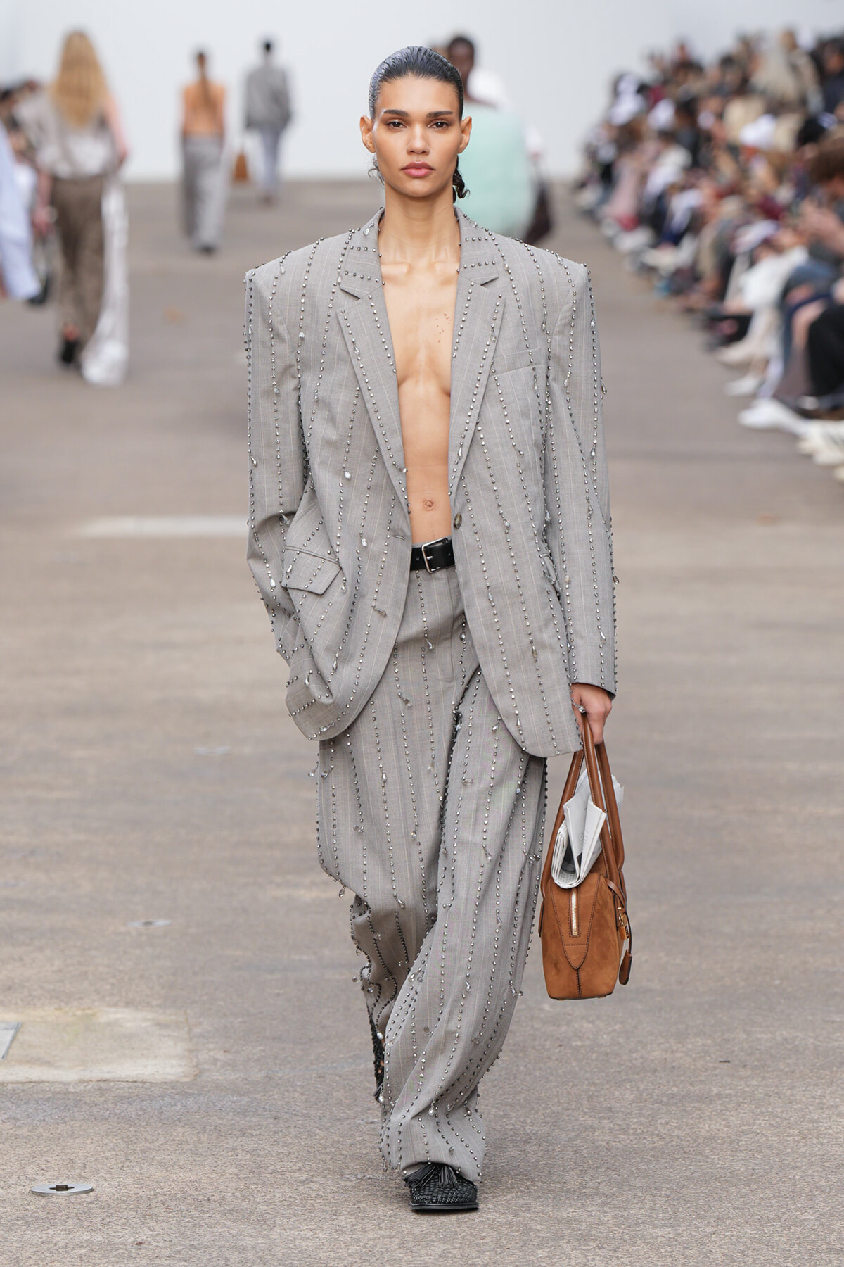 Model walking the runway in an oversized gray beaded pinstripe suit, blazer open and shirtless, carrying a brown tote with newspapers inside at the side.