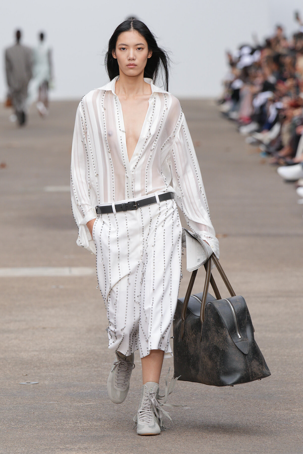 Model walking a runway in a sheer white striped blouse with studded trim, white pants, black belt, gray lace-up boots, and a large black handbag.