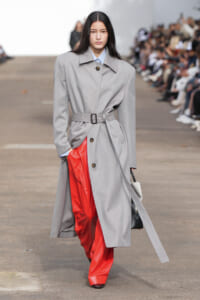 Model on a runway wearing a long gray belted trench over bright red pants, carrying a black-and-white handbag.