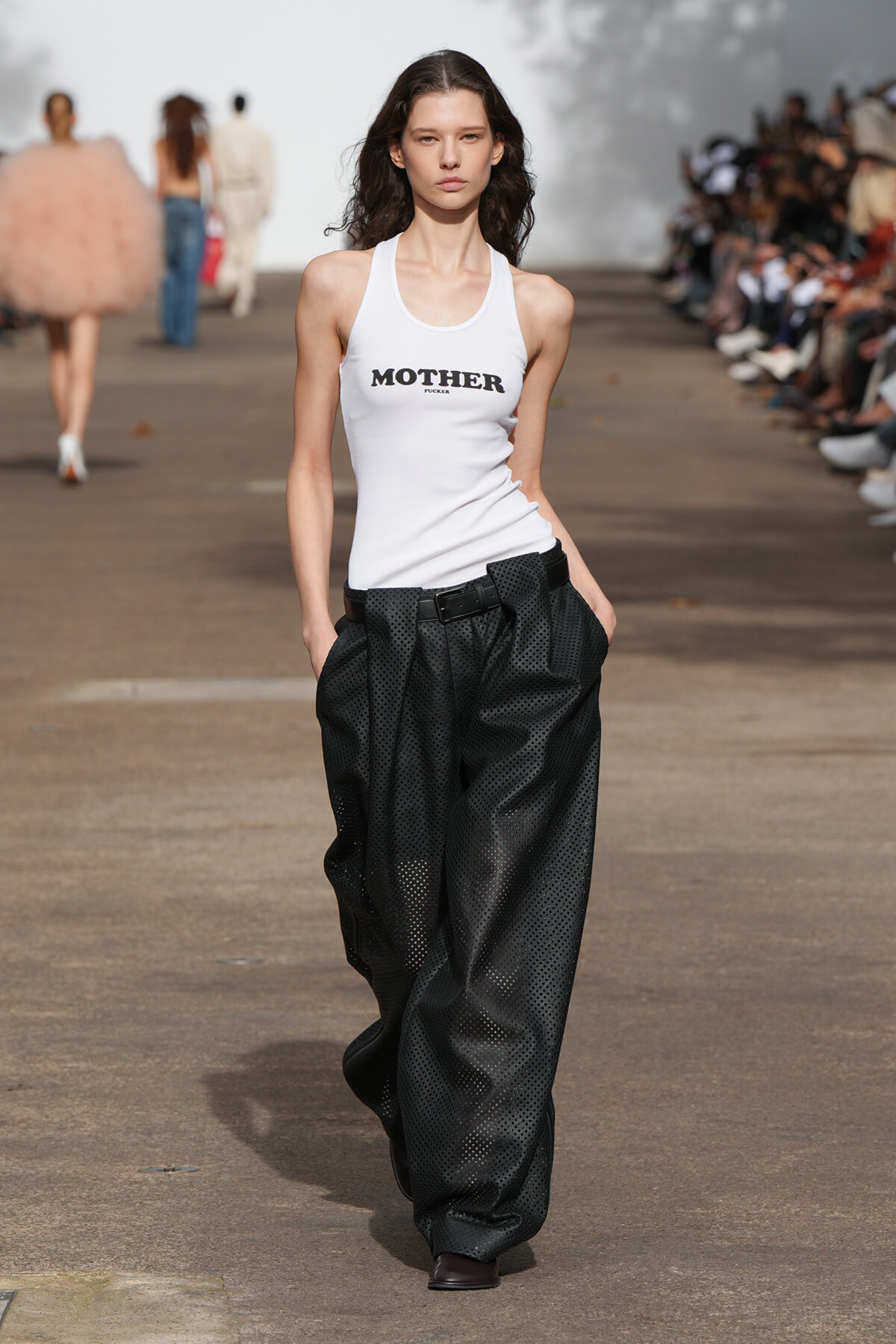 Model walks a runway wearing a white tank top with a bold 'MOTHER' logo and oversized perforated black pants, hands in pockets, audience on both sides.