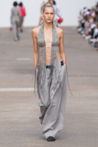 Model walks the runway in a silver fringe halter top and loose gray trousers, showcasing a metallic, fringe-heavy look.