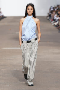 Model walking a runway in a pale blue halter-top paired with wide, sparkling silver trousers and a black belt.