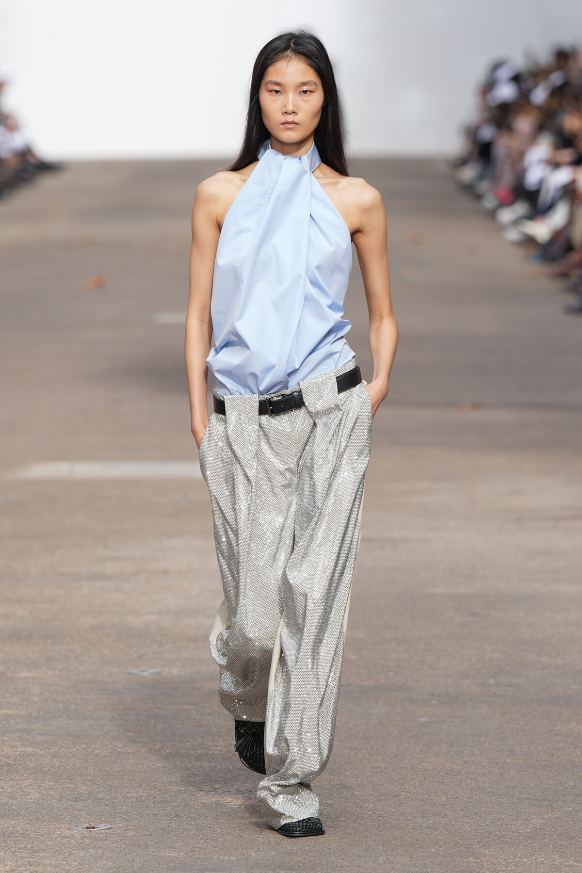 Model walking a runway in a pale blue halter-top paired with wide, sparkling silver trousers and a black belt.