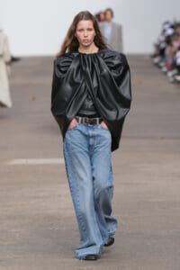 Model on a runway wearing an oversized black leather top with dramatic puff sleeves, paired with loose blue jeans and a brown belt.