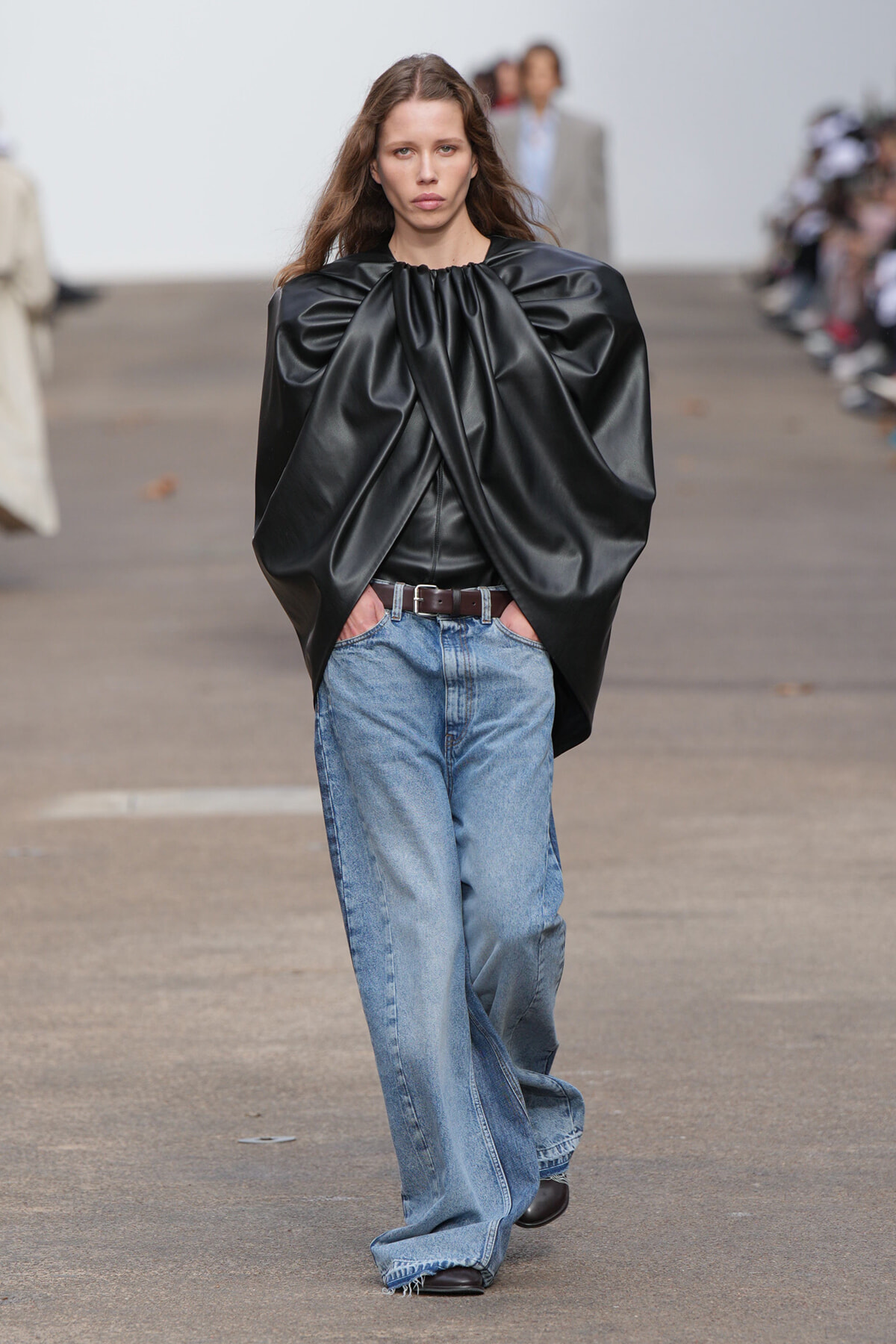 Model on a runway wearing an oversized black leather top with dramatic puff sleeves, paired with loose blue jeans and a brown belt.