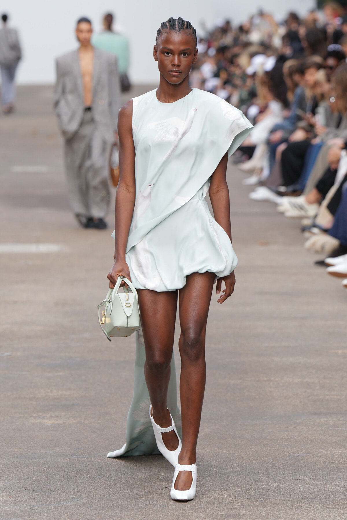 Model walking a fashion show in a pale bluish-green draped mini dress, carrying a small pale handbag and wearing white Mary Jane heels. Audience blurred in background.
