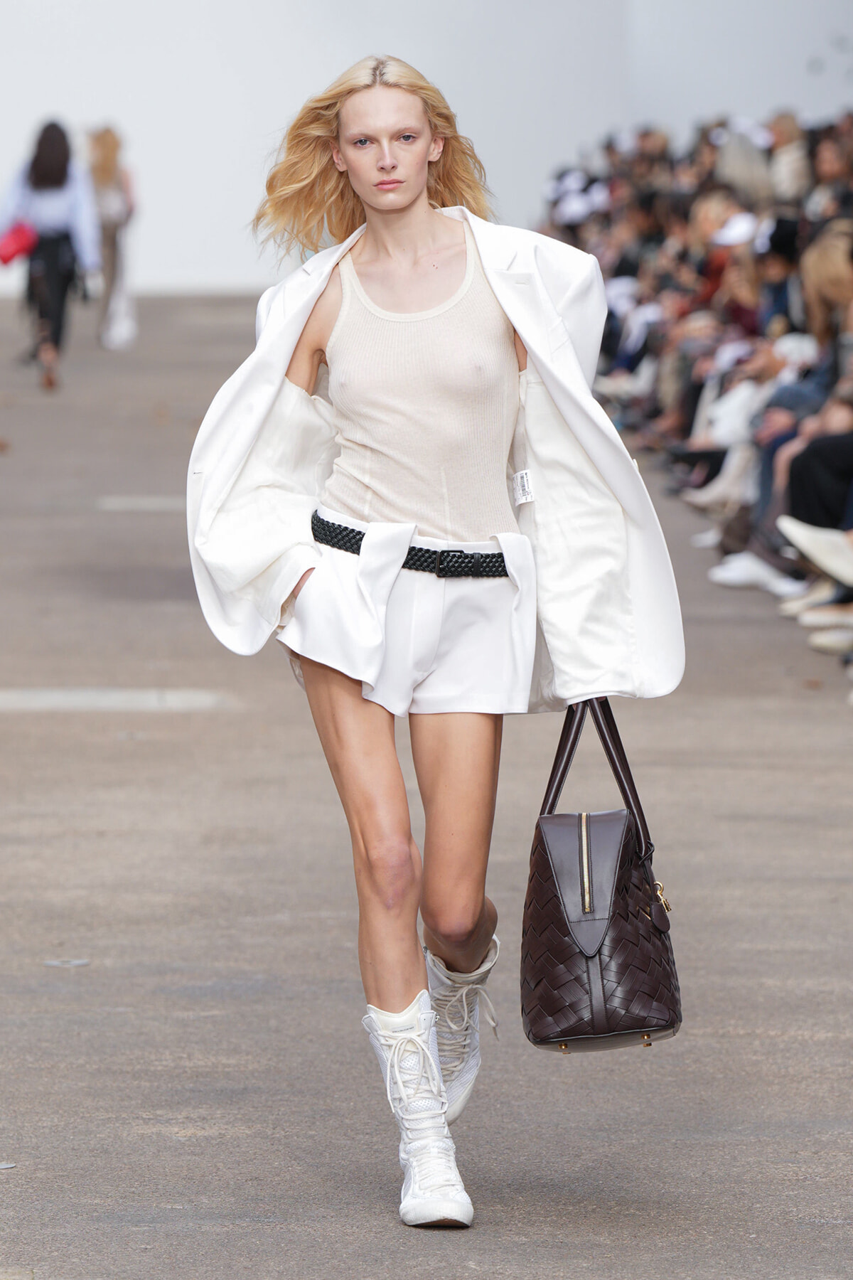 Model walks runway in white sleeveless top, white shorts, and oversized white blazer, carrying a woven brown bag and white knee-high boots.