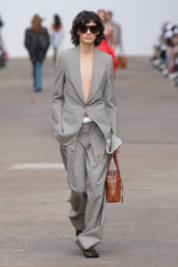 Runway model in a gray pinstripe suit with a deep-plunge blazer, oversized trousers, dark sunglasses, carrying a newspaper and a brown handbag.