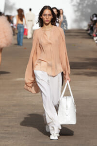 Model walking a fashion show runway in a peach sheer blouse with billowy sleeves, white wide-leg pants, white woven tote, white sneakers, and clear glasses.