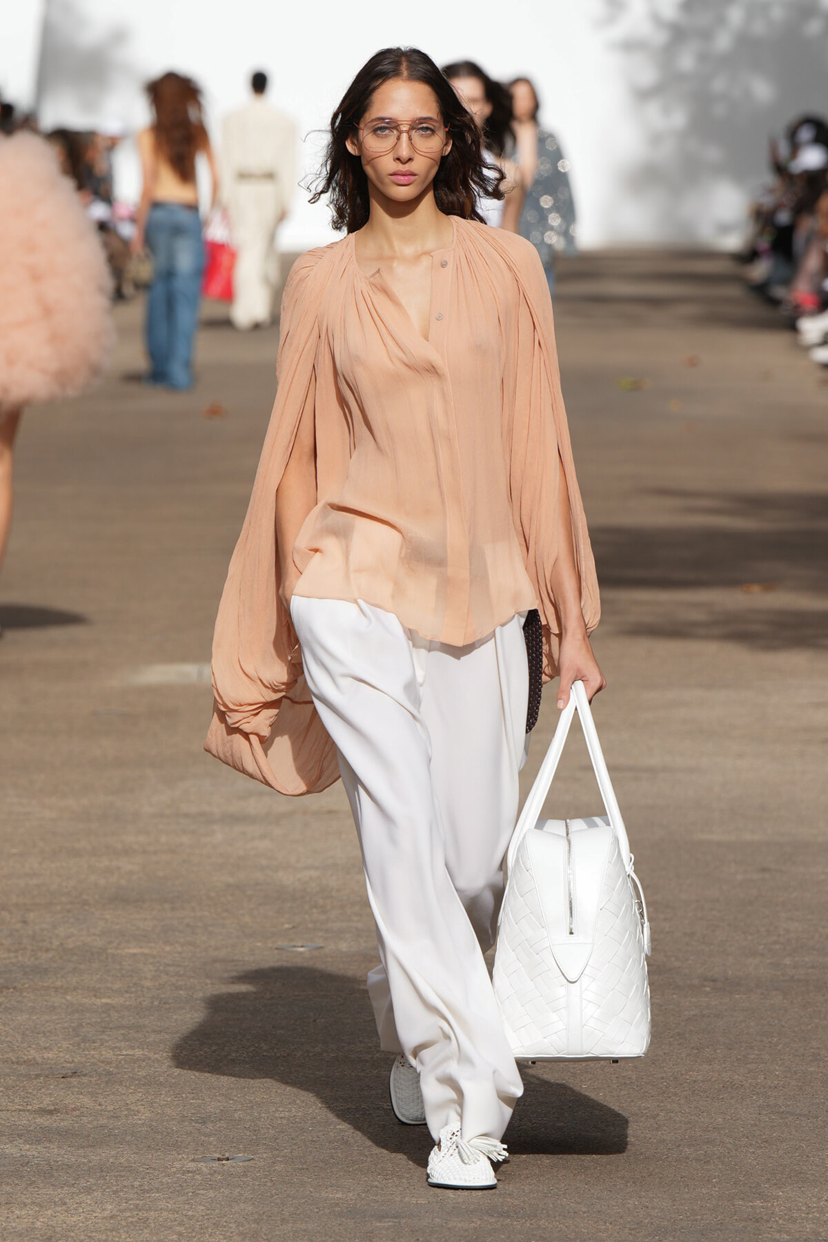 Model walking a fashion show runway in a peach sheer blouse with billowy sleeves, white wide-leg pants, white woven tote, white sneakers, and clear glasses.
