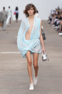 Model walks the runway in a light blue sheer draped dress with a deep side cutout, carrying a small silver handbag and wearing white lattice-heel sandals.