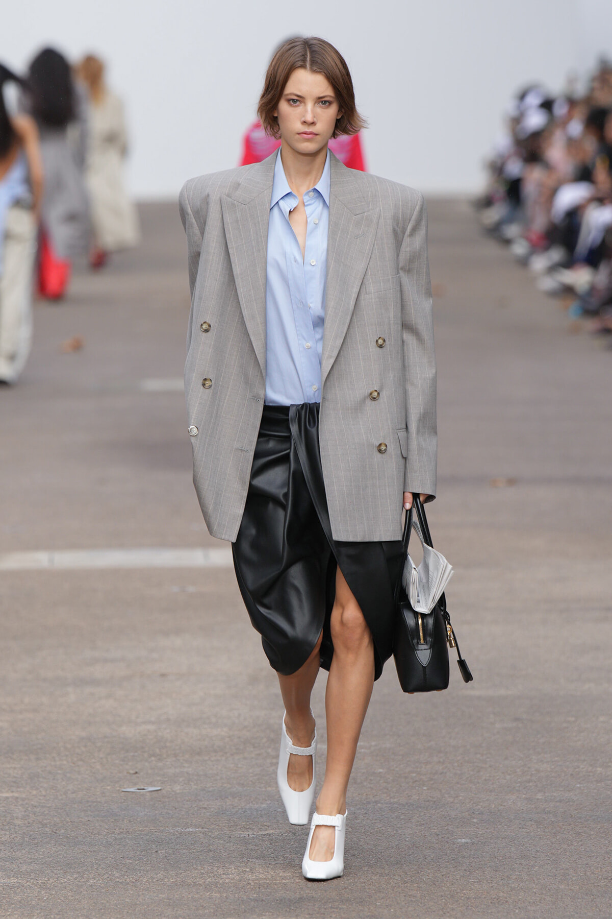 Model walking runway in a oversized gray pinstriped blazer over a light blue button-down, black draped skirt, white heels, and a black bag.