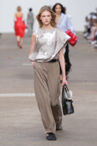 Model walking a runway in a taupe satin one-shoulder top with white bird prints and wide tan trousers, carrying a black handbag with newspapers peeking out.