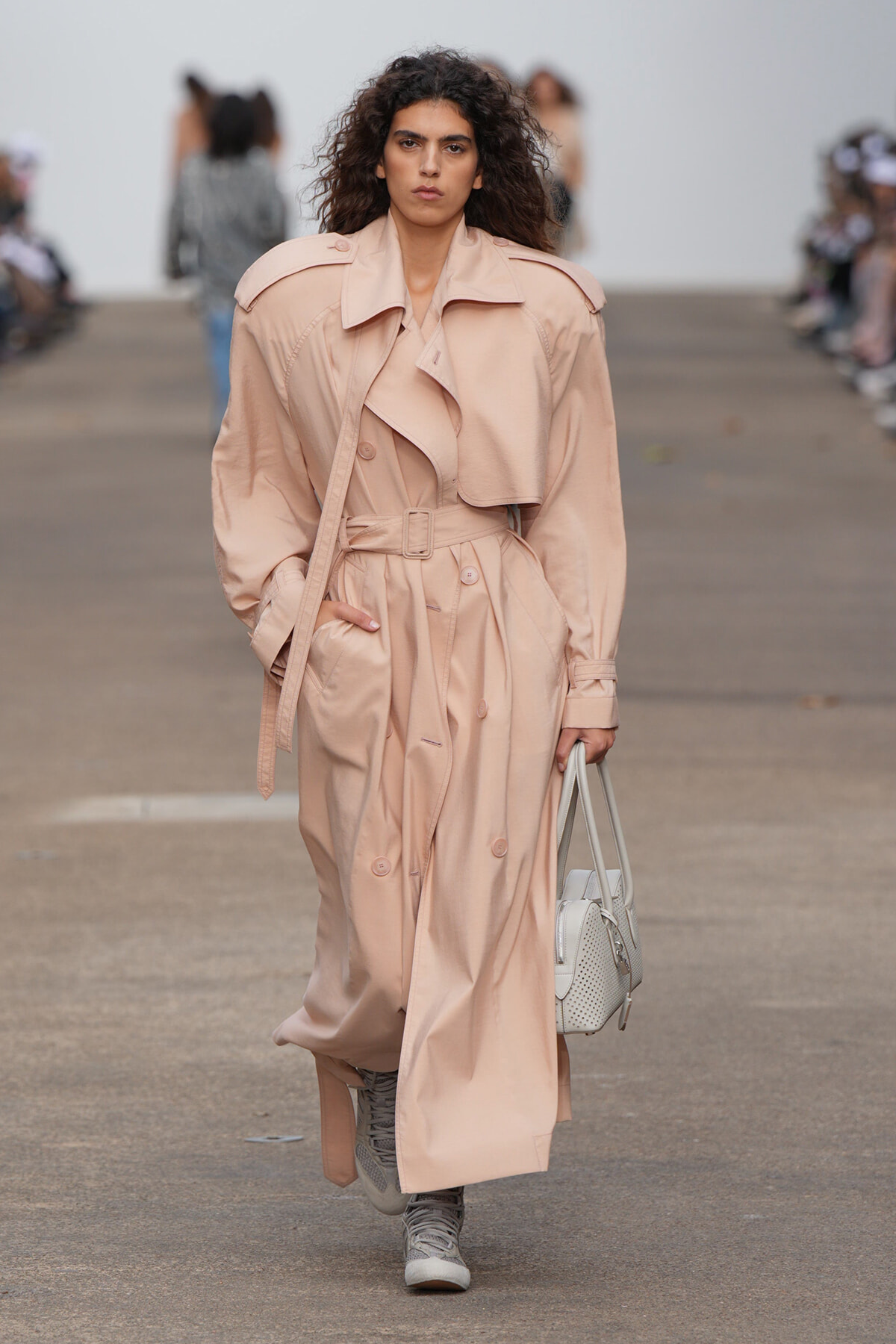 Female model walking a runway in a long blush trench coat with a belt, holding a light gray handbag. The background is a blurred audience.