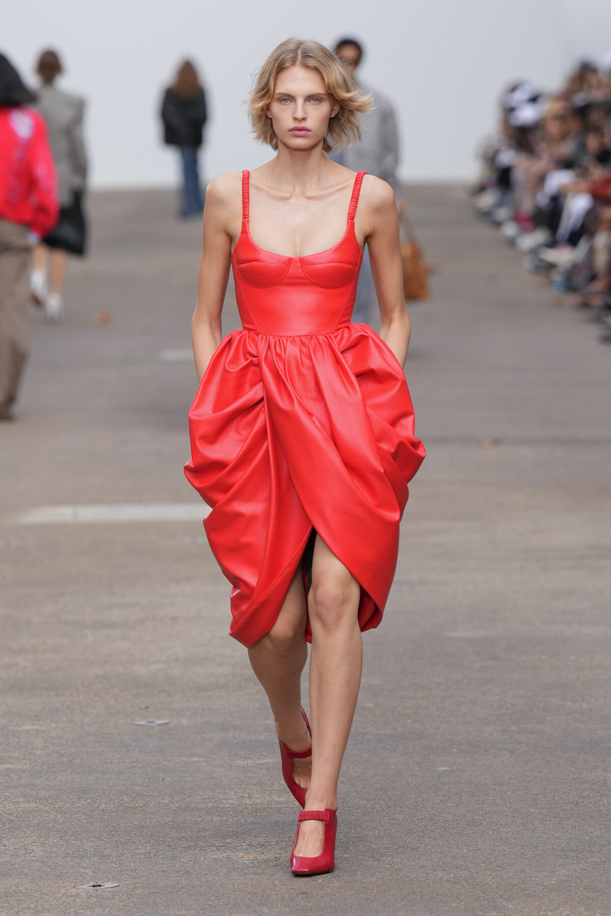 Female model wearing a bright red satin dress with a fitted bodice and voluminous draped skirt walking a runway.