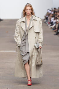 Model on a runway wearing a long beige trench coat over a gray suit with a draped blazer, carrying a small gray handbag and red heels.
