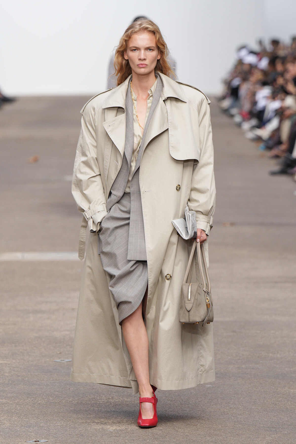 Model on a runway wearing a long beige trench coat over a gray suit with a draped blazer, carrying a small gray handbag and red heels.