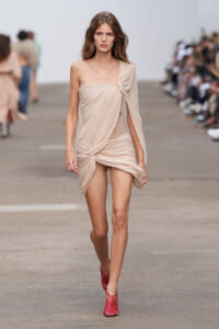 Model walks a runway in a beige one-shoulder, draped dress with twisted front details, paired with red woven-strap heels.